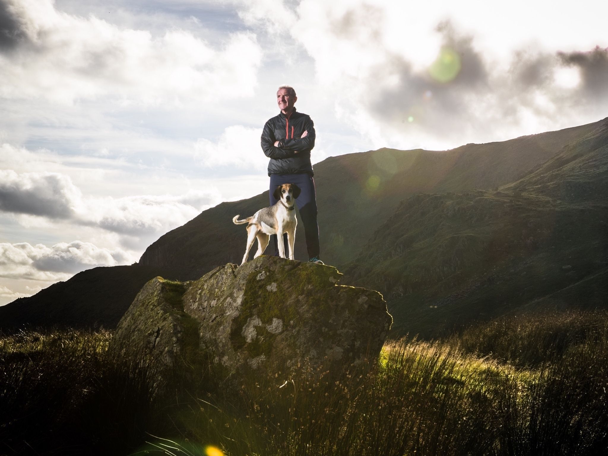 man standing beside a dog on a rock in front of a mountain